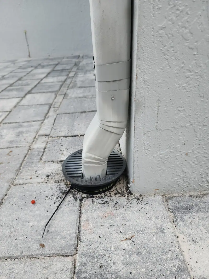 A white downspout running into a ground outlet on a paver patio